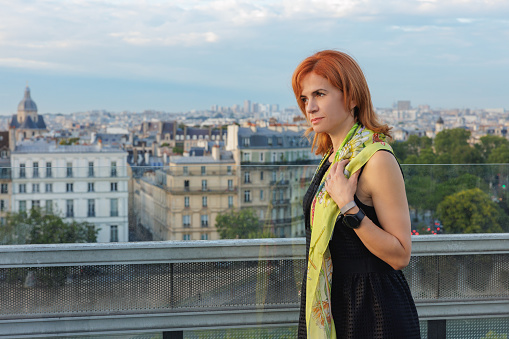 Beautiful mid adult businesswoman standing on top floor building terrace, leaning on the glass fence, Paris cityscape under the sky, modern business people
