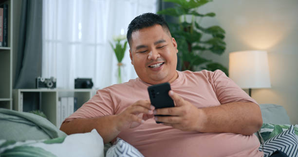 Chubby Man Using Smartphone to relaxing on sofa in Living Room Young happy man immersed in digital realm of living room. connection through a smartphone, where online networking and messaging provide moments of enjoyment. relaxation with every swipe and tap. asian man obese stock pictures, royalty-free photos & images