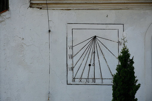A close up on an old solar clock with all its elements made out of metal seen on the wall of an old church or cathedral next to a big shrub growing next to it seen on a sunny summer day