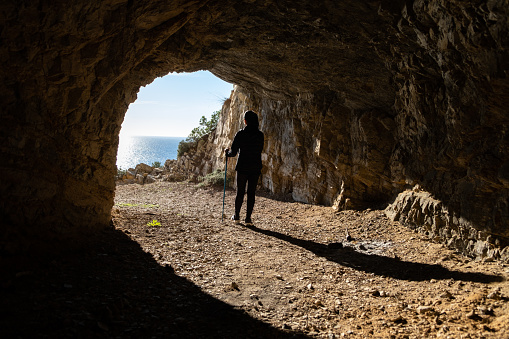 Unrecognizable woman with hiking poles looks relaxed at the exit of a cave on a beautiful bright day with the sea in the background on the Spanish Costa Brava