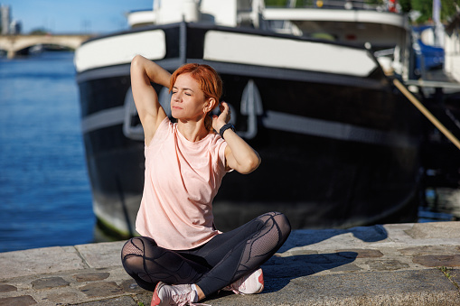 Beautiful mature woman sitting cross-legged and eyes closed on stone fence at Paris waterfront on a sunny day and stretching arms, healthy lifestyle and exercise