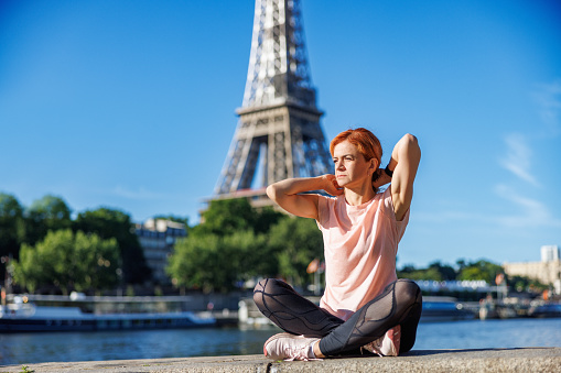 Beautiful mature woman sitting cross-legged on stone fence at Paris waterfront on a sunny day and stretching arms, Eiffel Tower in the background, healthy lifestyle and exercise