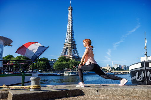 Beautiful mature woman stretching in lunge position on stone fence at Paris waterfront on a clear day, Eiffel Tower in the background, healthy lifestyle and exercise