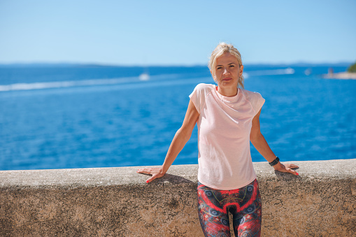 Portrait of beautiful middle aged woman wearing T-shirt and leggings leaning on stone wall on a sunny day, blue seascape and horizon under the clear sky in the background, enjoying her summer break