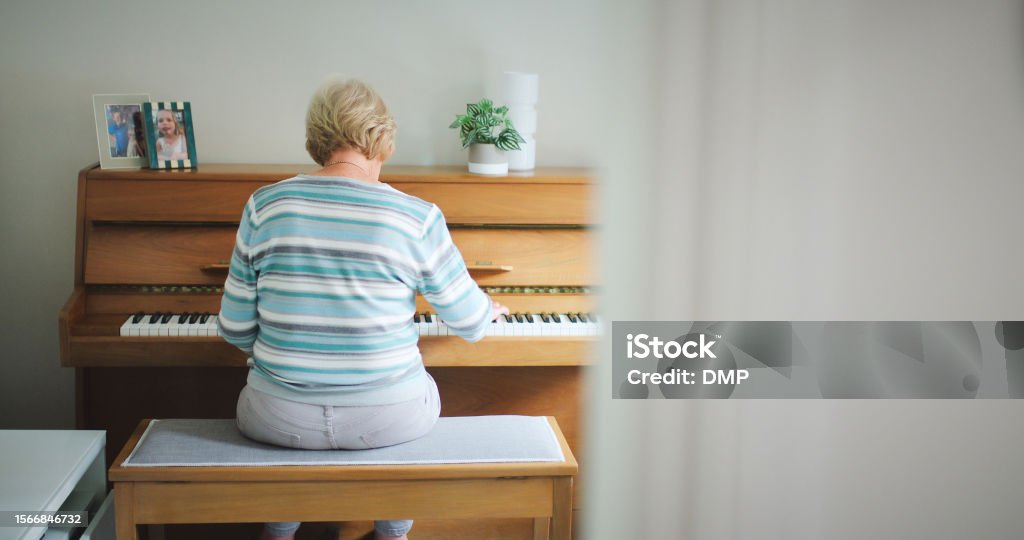 Piano, music and a woman playing at home for performance, hobby or to relax in a room. Behind senior female person with an old or vintage instrument with keys and notes on wooden bench in a house 80-89 Years Stock Photo
