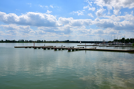 A close up on a set of wooden marinas, piers, or platforms located next to a sandy beach covered with grass located next to a vast lake or river with some boats swimming on it in summer