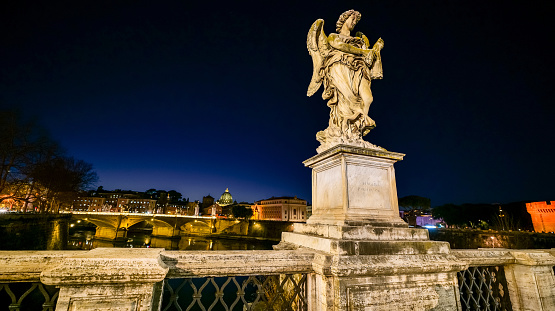 An idyllic twilight cityscape of the Baroque heart of Rome seen from Ponte Sant'Angelo, with a Bernini's angel in foreground. Along the horizon, the Vittorio Emanuele II birdge and the silhouette of the majestic dome of St. Peter's Basilica. Initially this bridge was built under the emperor Hadrian in 134 AD. to connect the heart of Rome with his mausoleum, currently Castel Sant'Angelo. A few centuries later, under the pontificate of Pope Clement IX, the architect and sculptor Giovanni Lorenzo Bernini was commissioned to create the current balustrade of the bridge, decorated with ten statues of the Angels of the Passion of Christ. According to tradition, it was Bernini's pupils who sculpted the statues of the Angels in 1669, converted Ponte Sant'Angelo in a Baroque masterpiece. In 1980 the historic center of Rome was declared a World Heritage Site by Unesco. Super wide angle image in 16:9 ratio and high definition quality.