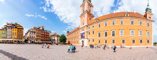 The center of Warsaw's Old Town, providing a view of Castle Square and Warsaw Castle. People are walking along the square, and the sky above appears blue with scattered clouds.