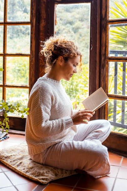 Knowledge for all healthy natural midnful zenlike lifestyle people. One adult woman sitting on the floor at hoe in total relaxation indoor leisure activity alone reading a book. Nature outside view stock photo