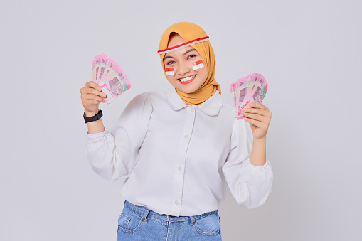 Smiling young Asian Muslim woman wearing hijab Celebrate Indonesian independence day on 17 August while holding money Indonesian rupiah banknotes isolated over white background