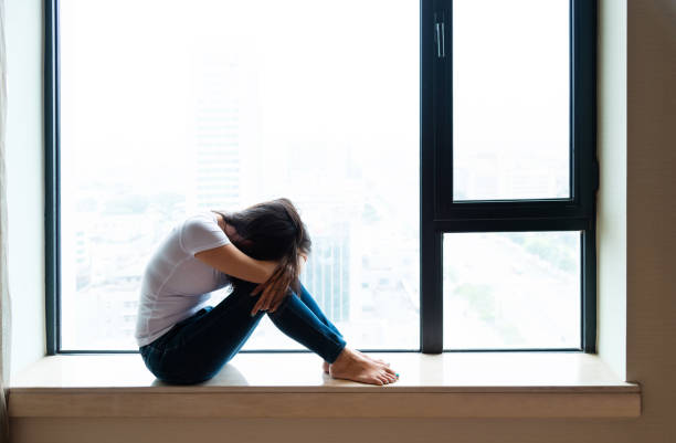 istock Sad woman sitting on window sill 1566902848