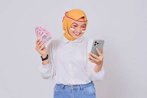 Cheerful young Asian Muslim woman wearing a hijab holding money and mobile phone isolated over white background. Celebrate Indonesian independence day on 17 August