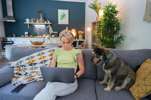 Young Caucasian woman using a laptop, while her mixed-breed dog makes her company on the sofa