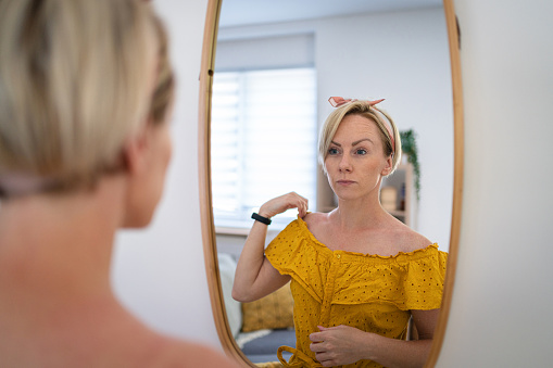 Caucasian woman in a yellow dress observing herself in the mirror while adjusting her dress