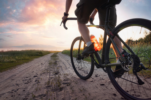 Man riding a gravel bicycle on the trail at sunset. Colorful landscape with cyclist, bike, field, green grass, dramatic sky with bright sunlight. Sport and travel. Low angle view.