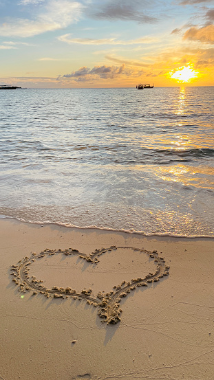 The figurine of the heart is in the sand on a luxurious sandy beach. Footprints in the sand. Waves in the sea. Dawn in the sky. Tropical layout for Valentine's Day.