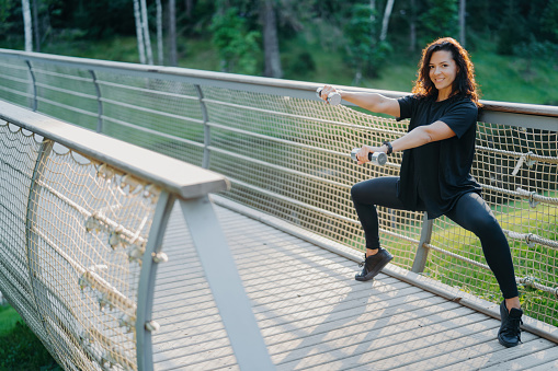 Active woman does squats with dumbbells at a bridge, wearing active wear, training biceps with sport equipment. Expresses joy in sporty lifestyle concept.