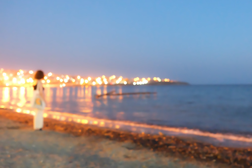 Out of focus coastal city in night time, woman standing at the beach, thoughtfully watching city lights