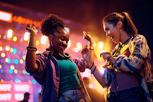 Happy African American woman and her female friend having fun and dancing while attending open air music concert at night.