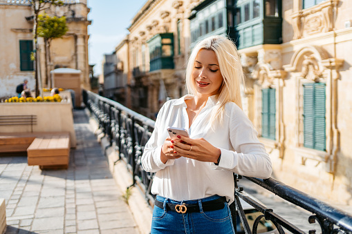 Beautiful young blonde woman texting on her smart phone while leaning on a railing in Valletta, Malta.
