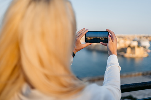 Beautiful young blonde woman taking pictures of the view using her smart phone from a high viewpoint in Valletta, Malta.