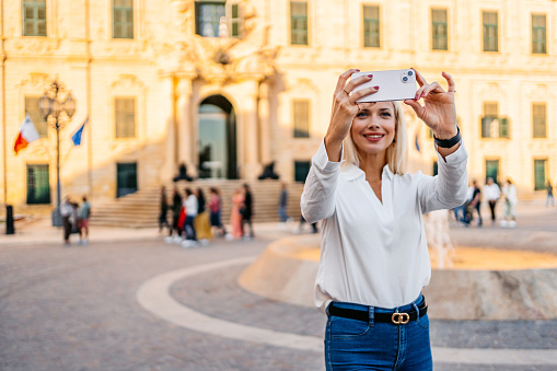 Beautiful young blonde woman taking pictures using smart phone of the view from the town square in Valletta, Malta.
