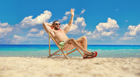Young man in swimwear sitting on a beach chair by the sea and waving