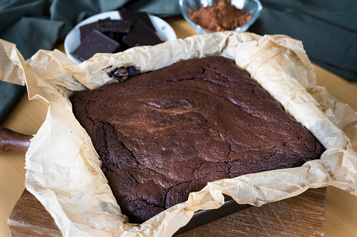 Chocolate brownie cake baked in a baking tin with parchment paper in the baking tin and cocoa powder and chocolate in the background.