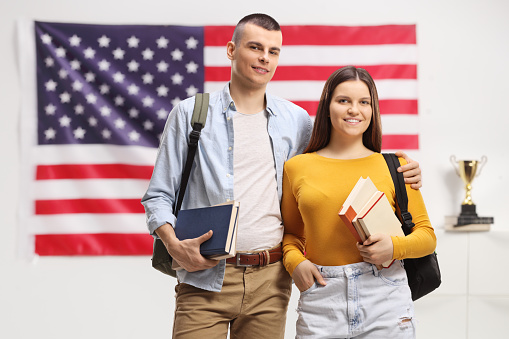 Male and female students with backpacks and books standing in front of an American flag