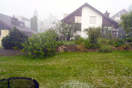 Heavy summer storm with hail falling down. There are hailstones on the lawn and the visibility is strongly reduced due to the dense rain and strong wind.