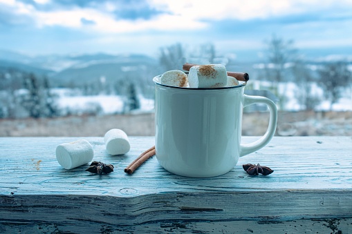 Christmas, New Year, a cup of hot coffee with marshmallows and spices, Christmas decorations, against the backdrop of a winter landscape, on a wooden table