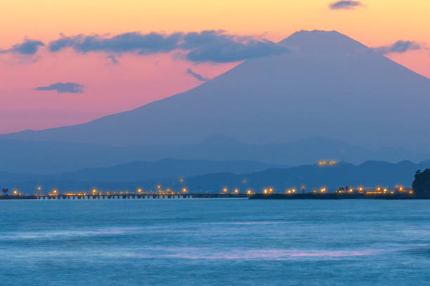 Enoshima Coast,Inamuragasaki, Kamakura, Kanagawa ,Japan-March 8 ,2023: a peaceful twilight sky with a mist-shrouded Mount Fuji in the distant backdrop.a serene twilight view from the coast near Inamuragasaki and Enoshima in Fujisawa City, Kanagawa Prefecture, Japan. As the sun sets, the striking silhouette of the iconic Mount Fuji stands tall on the horizon, its top enshrouded by a layer of misty haze, enhancing its mystic allure. Dominating the foreground, a male skateboarder is caught in action, confidently gliding across the coastal path, while another man, slightly blurred in the distance, is seen deftly surfing through the ocean waves. The scene is perfectly captured with a calm lake in the middle ground, its mirror-like surface reflecting the majestic mountainous terrain and the subtle pastel hues of the twilight sky. The entire composition blends harmoniously to create a truly tranquil and picturesque environment, encapsulating the unique beauty of a Japanese coastal dusk.