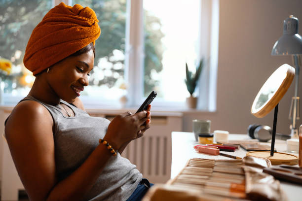 istock Woman of Black ethnicity using mobile phone while doing her make up 1567310951