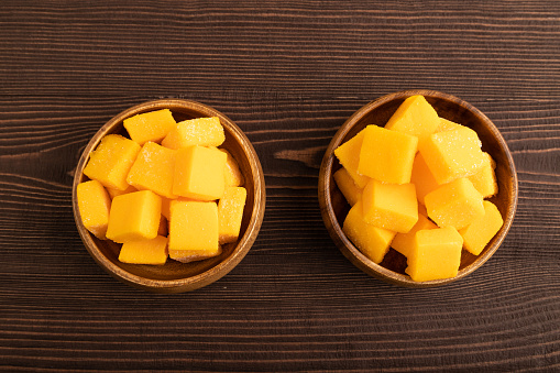 Dried and candied mango cubes in wooden bowls on brown wooden textured background. Top view, flat lay, close up, vegan, vegetarian food concept.