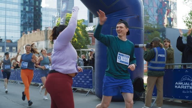 Slow Motion Portrait of a Smiling Plus Size Female Runner Crossing the Finish Line, Celebrating and Demonstrating her Willpower. Friendly City Marathon Participants Waving at the Audience