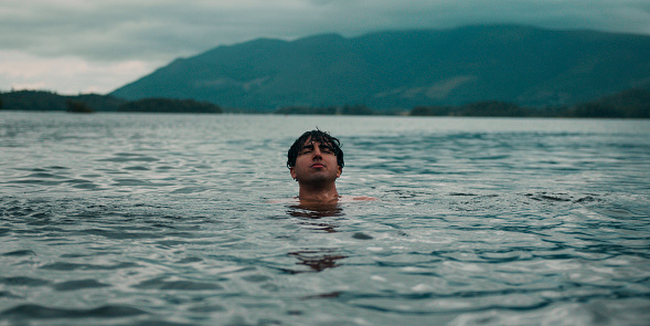 An eye level of a teenage boy wild swimming in a lake in Keswick, the Lakes. He is enjoying the cold water dip, closing his eyes tipping his head back. He smiles at the camera, swimming closer to it.