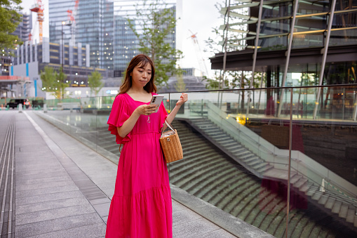 Woman in pink dress walking in urban city, using smart phone