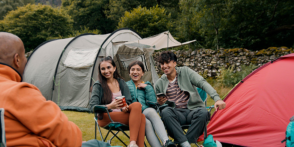 Over the shoulder view of a small group of siblings sitting together on camping chairs smiling and using a smartphone, they are camping and have set up some tents to create a campsite at Hollows Farm, The Lake District in Cumbria, England. They are sitting with their father.