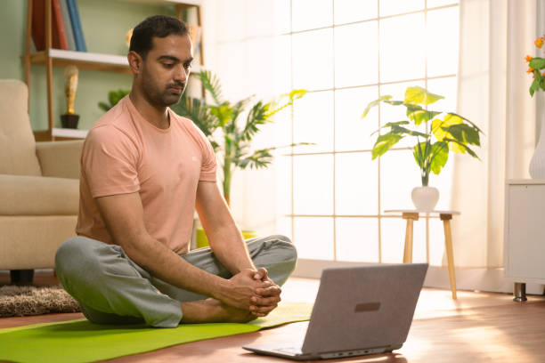 istock Indian man doing exercise by watching yoga video tutorial or online class from laptop at home - concept of virtual fitness training, stretching workout and Digital Wellness 1567402880