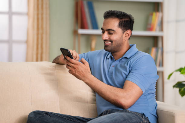 istock Happy relaxed indian man using mobile phone while sitting on sofa at home during Sunday - concept of online dating, relaxation and social media 1567405787