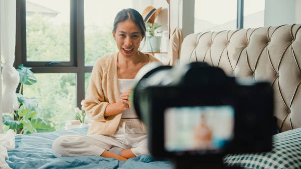 Happy Asian girl influencer with white cream pajamas recording sharing morning routine in camera sit on fluffy comfort bed talk with subscriber in bedroom morning light. Female morning vibes. stock photo