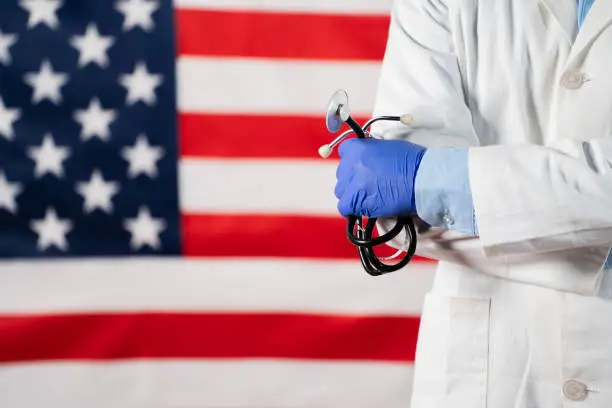 Confident Doctor standing by holding stethoscope in hand in front of US flag - concept of US labour day celebration showing with copy space.