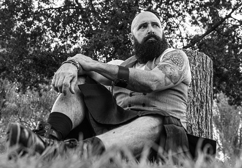 low angle shot of a mature bearded man with shaved head dressed in skirt sitting on the grass leaning his back on a tree stump and llooking something far away
