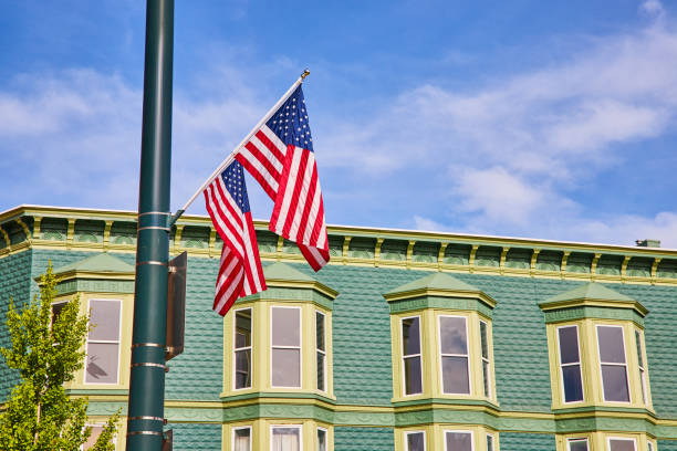 Image of Two American flags on pole with blue sky and a green tree in front of teal brick building