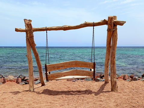 Rustic swing on the beach sand, sea and turquoise water under blue sky of the Dahab desert. Wooden and rope swing. Games and outdoor fun.