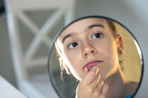 A teenage girl looks thoughtfully at her reflection in the mirror, the concept of changes in the body in adolescence, the maturation period.