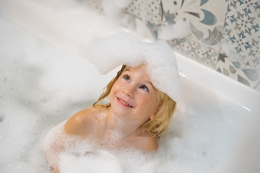 Cute funny caucasian blonde three years old baby girl taking bath with white foam bubbles,child playing,smiling ,having fun.Carefree childhood, kid body care concept.