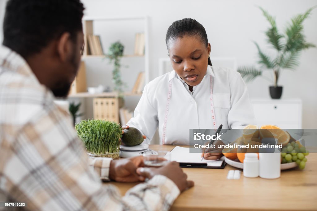 Expert in diet writing down details of meals for patient Focused female specialist writing details about fruit nutrients on clipboard paper while counselling client. Multicultural expert in diet formulating special meals for male patient in clinic. Nutritionist Stock Photo