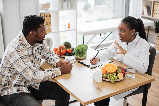 Attractive multiethnic lady in white coat having conversation with bearded man in casual wear indoors. Efficient expert in diet giving recommendation on use of food and nutrition in consulting room.
