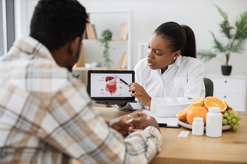 African american specialist in nutrition holding tablet with gastrointestinal tract on screen while counseling man at work. Focused woman explaining client about food intolerance during appointment.